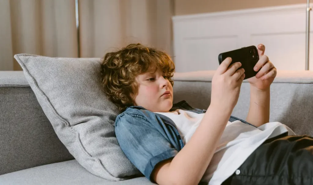 A young boy with curly hair is lying on a gray couch, propped up by a pillow, while intently looking at a smartphone in his hands. The warm indoor setting suggests a relaxed environment, but his deep focus on the screen highlights the growing concern about excessive screen usage among children. This image emphasizes the importance of screen time limits for children to encourage a balanced lifestyle that includes physical activity, social interaction, and offline learning.